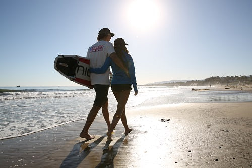 A couple enjoying Summer vibes while walking on the beach during sunset, holding a surfboard.