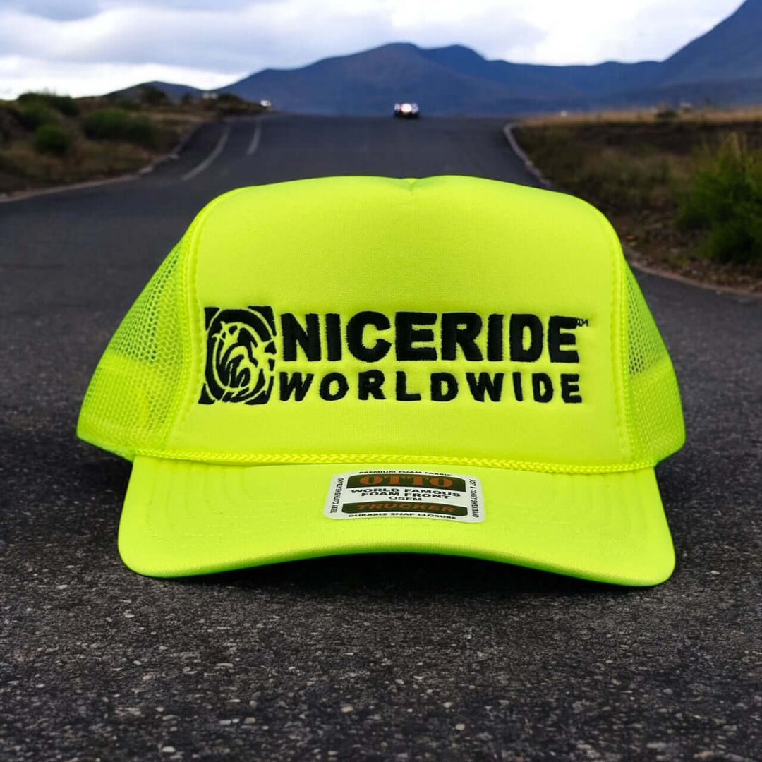 NICERIDE Worldwide bright yellow foam trucker hat on a road with mountains in the background.
