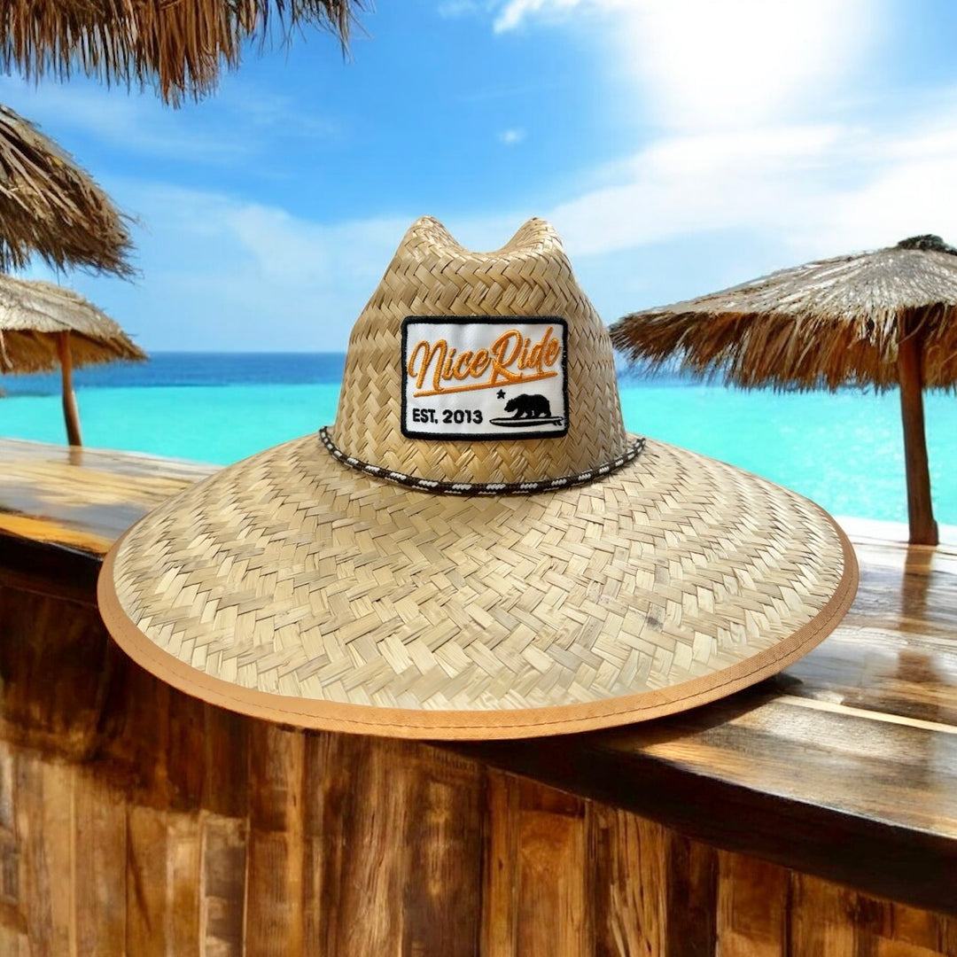 NICERIDE Big Sky Straw Sun Hat in natural straw, wide-brim design, showcased on a beachside bar with ocean backdrop.