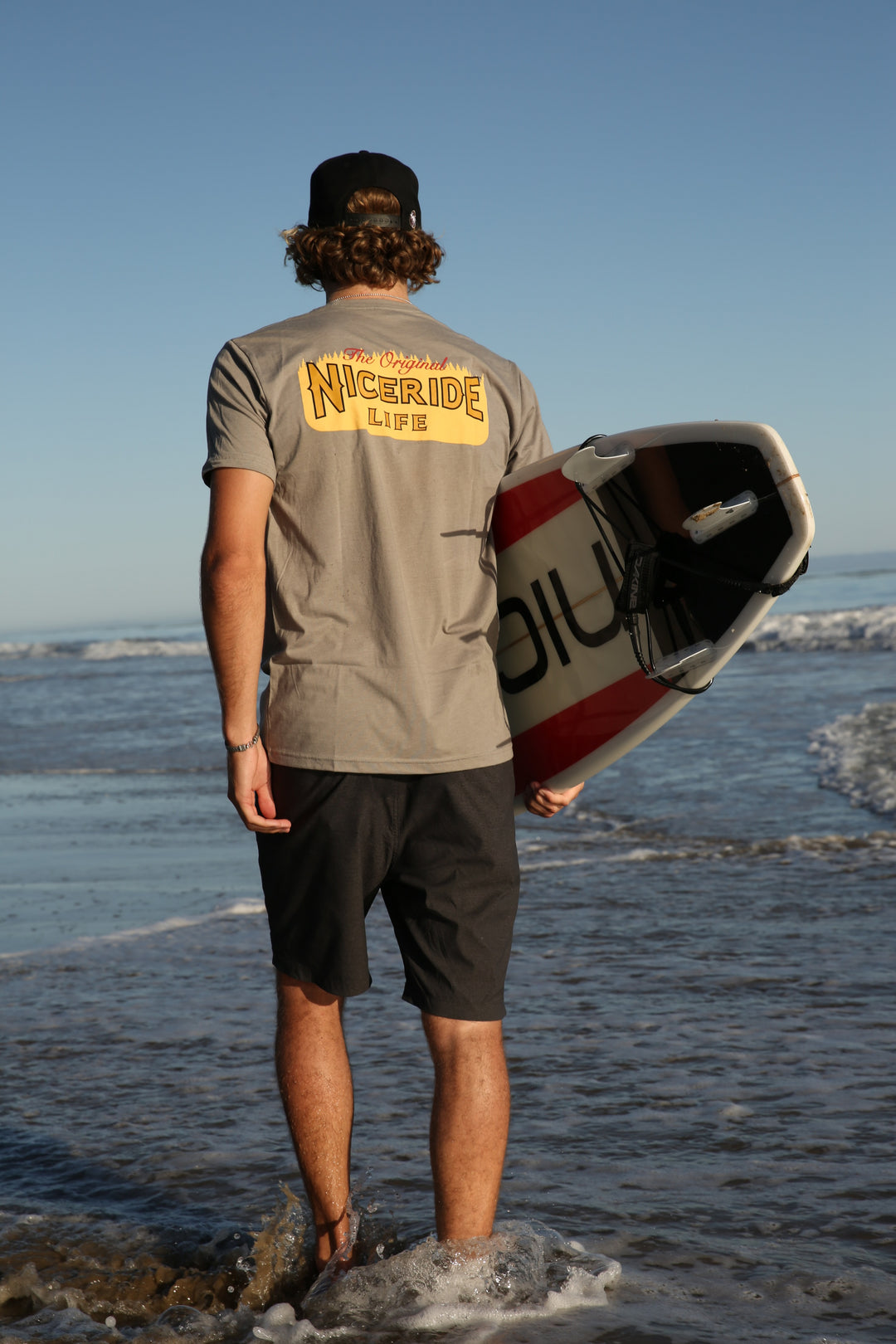 Man wearing a Next Level shirt at the beach holding a surfboard, showcasing NICERIDE design on back.