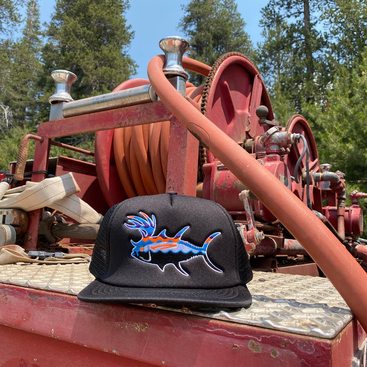 Black snapback trucker hat with embroidered fish design resting on a red fishing gear background in a natural setting.