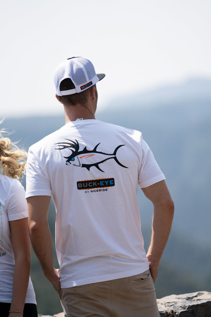 Man wearing a BUCK-EYE shirt with fishing design on back, standing outdoors in a scenic view.