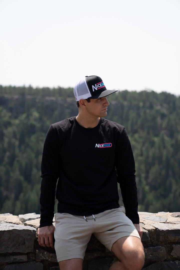 Man wearing The Tube black shirt and hat, sitting on a stone wall with a forest backdrop.