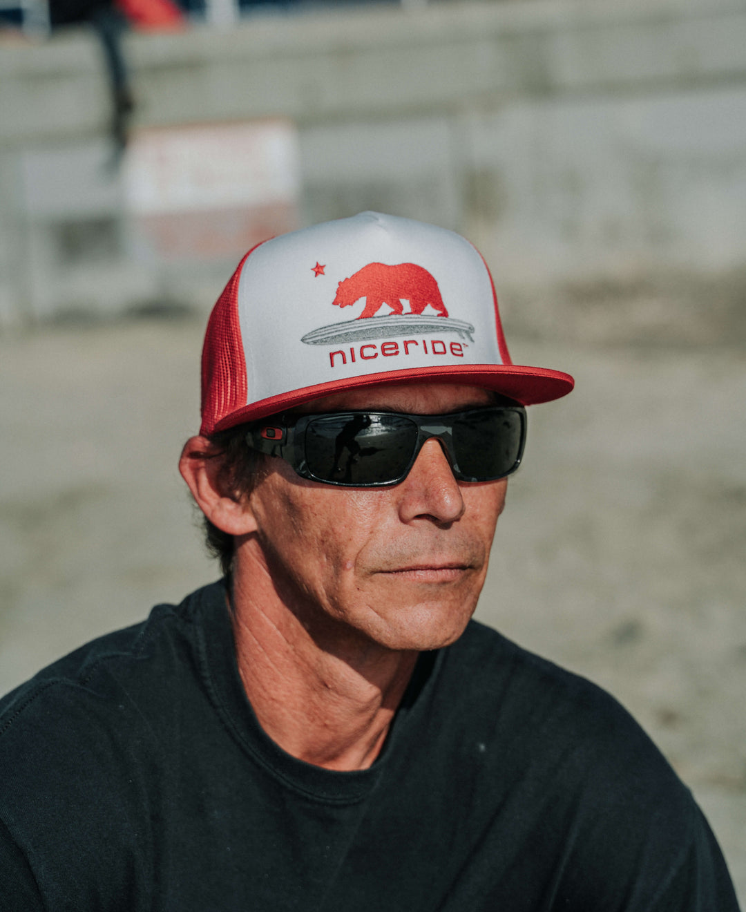 Man wearing a red and white "Hang 20" Trucker Hat by NICERIDE, featuring sunglasses and coastal background.