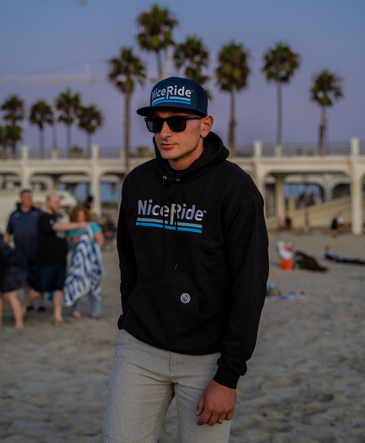 Person wearing a NiceRide hoodie and a hat at the beach, showcasing the Low Tide trucker hat.