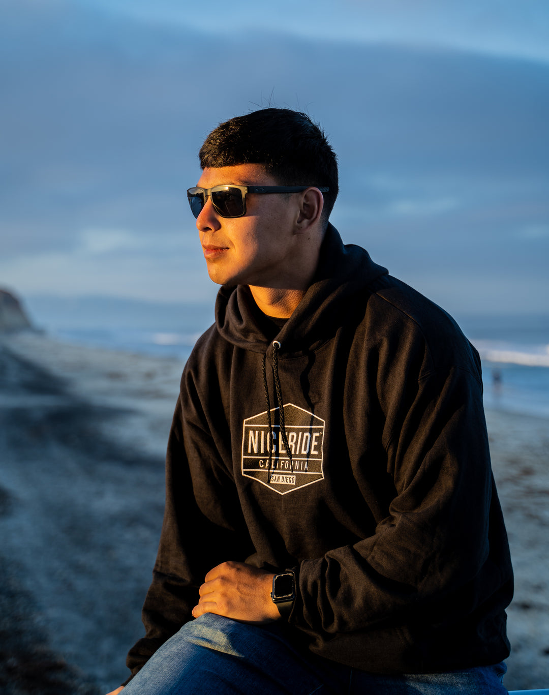 Man wearing a black hoodie with NICERIDE design at the beach during sunset.