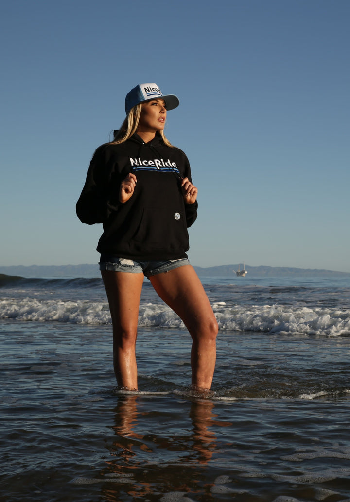 Woman wearing a black hoodie with NICERIDE logo standing in water, showcasing a stylish hoodie by the beach.